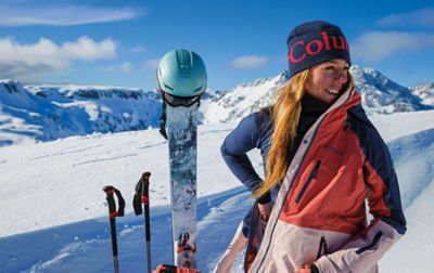 A skier stands on a snow-covered mountain taking off her ski jacket as her skis, poles, helmet, and backpack stick out of the snow next to her. Underneath her ski coat she is wearing a warm Columbia Sportswear baselayer. 