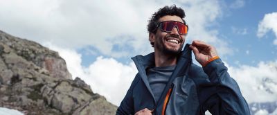 A man stands on a rocky mountaintop smiling widely as he unzips a blue Columbia Sportswear midlayer to reveal a baselayer underneath. 