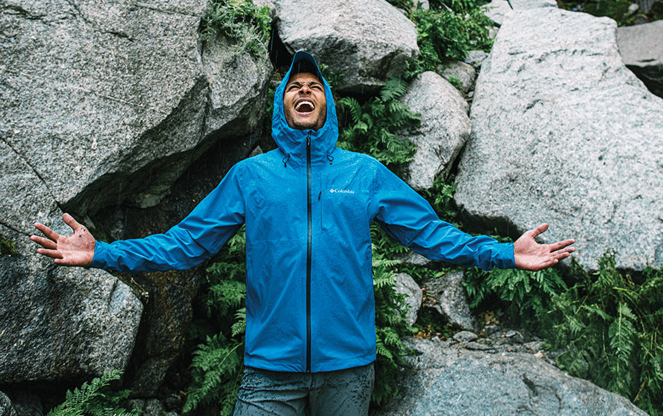 A man in a blue Columbia Sportswear rain jacket extends his arms out to each side and shouts jubilantly with rocks and ferns in the background.