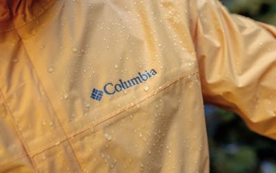 A close-up photo of the Columbia Sportswear logo on a yellow rain jacket with droplets of water all over it.