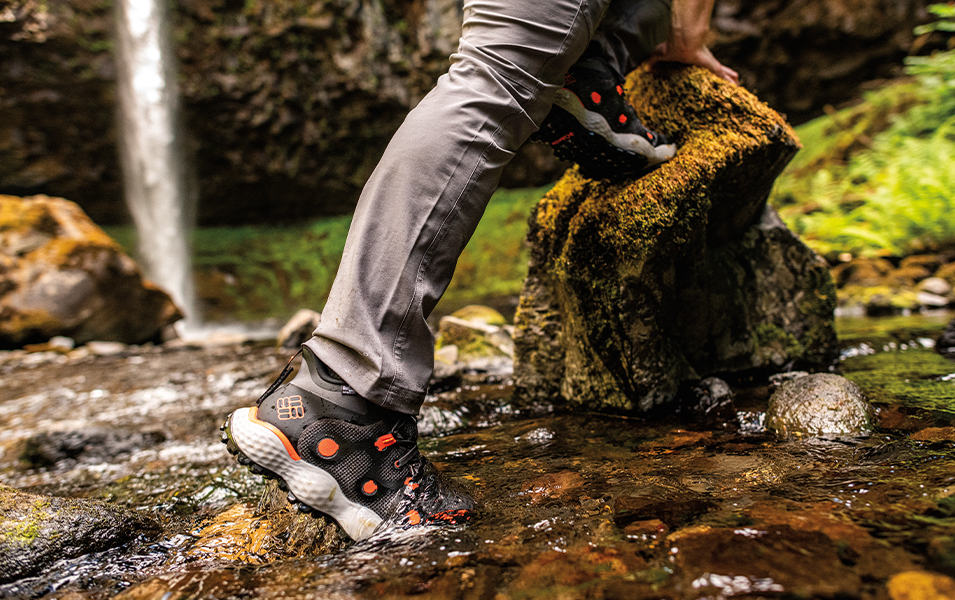 A close-up photo of a man’s legs in gray pants and black hiking shoes crossing a river with moss-covered rocks.