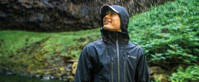 A man in a black Columbia Sportswear jacket leans to the side smiling in the rain with lush green foliage in the background.