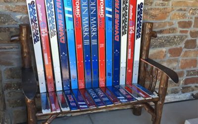 A patio bench made from colorful skis sits outside in a concrete area in front of a brick wall. 