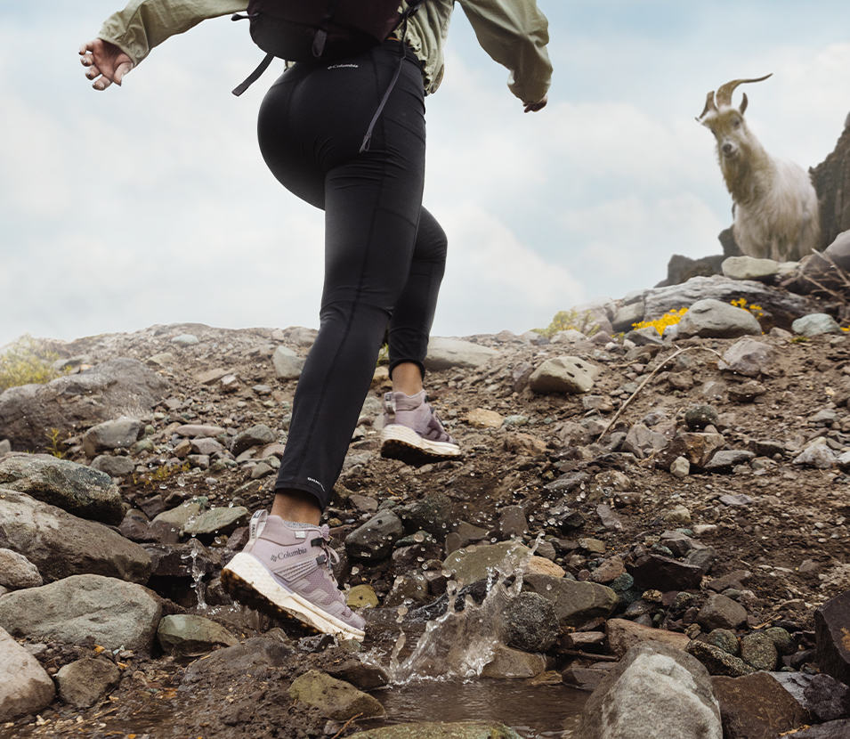 A man hikes up a rocky trail wearing Columbia Sportswear’s Facet 75 Mid shoe as a mountain goat watches in the distance. 