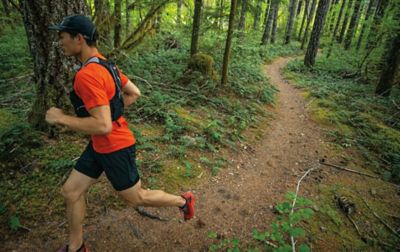A man in a red jacket and a black vest runs on a trail in a scenic forest area. 