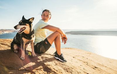 A woman sits on a rocky overlook above a sunny blue body of water with a smiling black dog at her side.