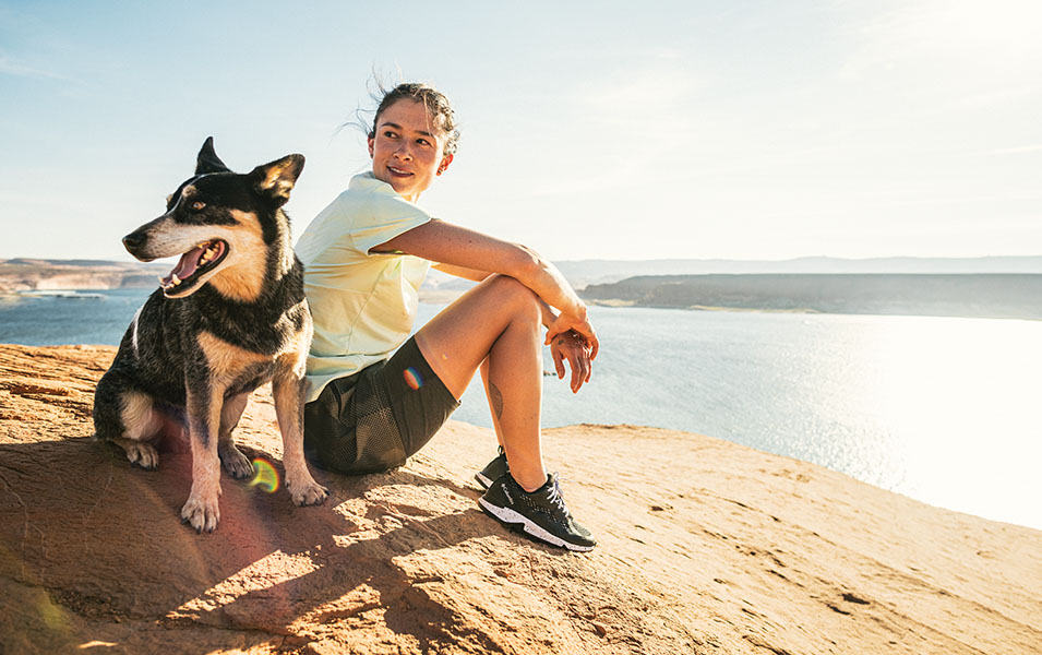 A woman sits on a rocky overlook above a sunny blue body of water with a smiling black dog at her side.