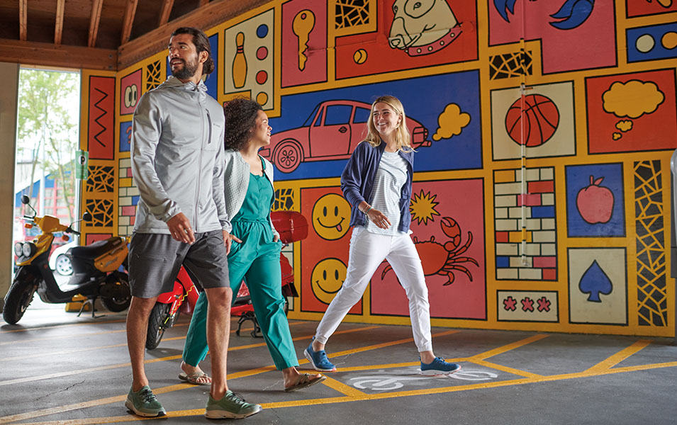Three smiling friends walk together through an outdoor parking lot with a colorful mural on the wall behind them. 