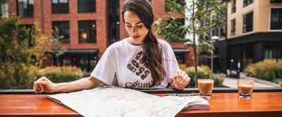A woman wearing a white shirt sits at an outdoor table in a metropolitan area looking at an adventure map with two cups of coffee beside her. 