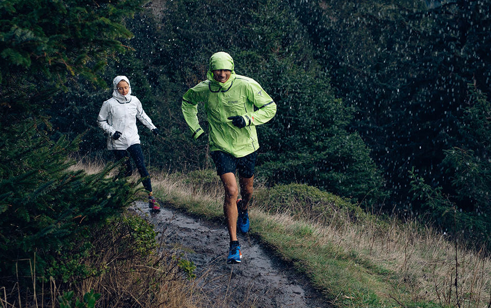 Yassine Diboun runs on a trail in the rain wearing a Columbia Sportswear raincoat with a woman in a white jacket behind him.