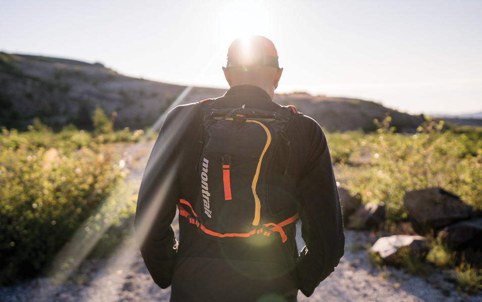 Yassine Diboun stands with his back to the camera on a sunny trail wearing a black running jacket and backpack.
