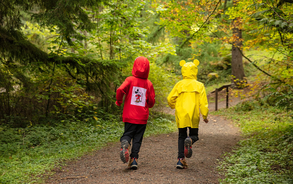 Two kids in rain coats running on a dirt path. 