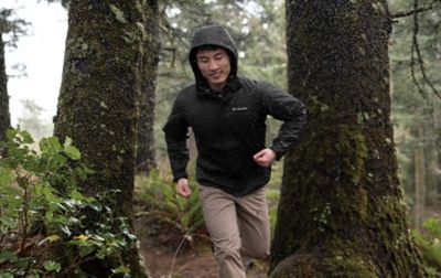 Homme courant sur un sentier forestier pluvieux portant une veste de pluie noire Columbia Sportswear.