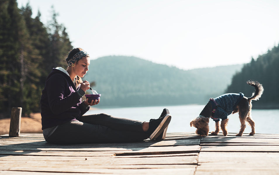 A woman sits on a dock eating food next to her dog with a picturesque body of water behind them.  