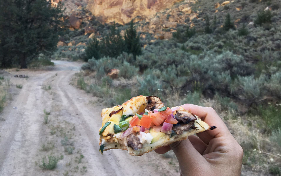 A hiker’s hand holds up a slice of pizza in front of a sprawling dirt trail. 