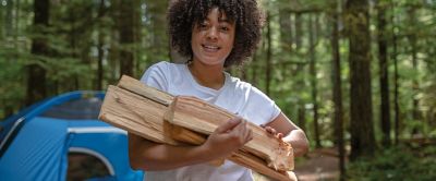 A woman with curly hairs holds a pile of firewood with a forest campground behind her.