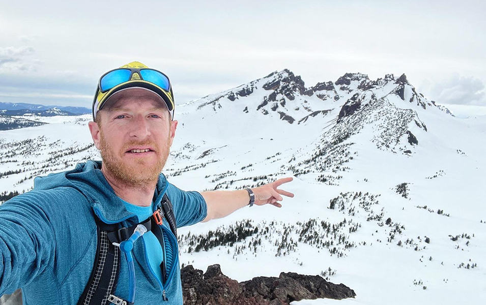 Professional trail runner Willie McBride takes a selfie on a mountain with a rugged snowy landscape in the background. 