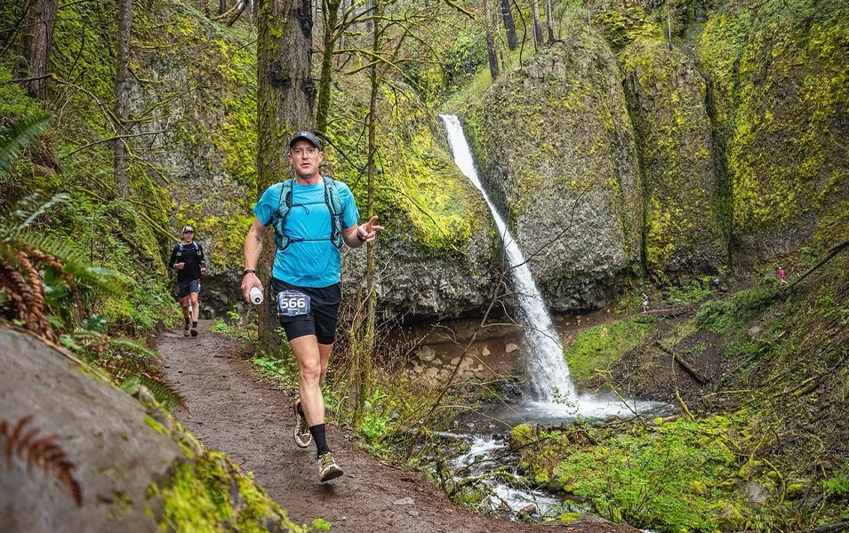 Professional trail runner Willie McBride runs through a mossy forest during an endurance race and smiles for the camera.

 
