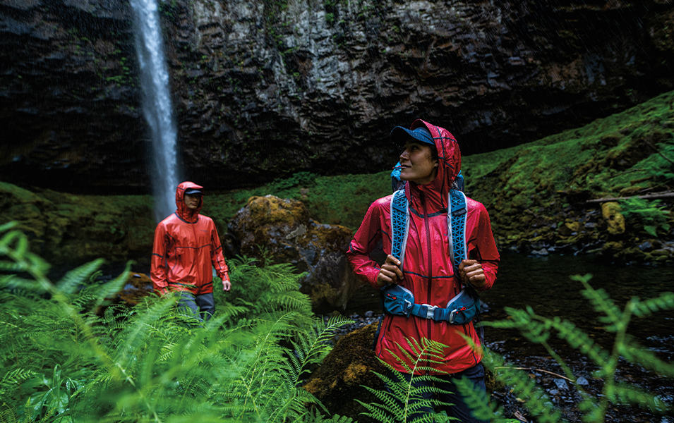 Two hikers wearing orange and red Columbia Sportswear OutDry™ Extreme rain jackets walk in front of a beautiful waterfall with lush green ferns around them.