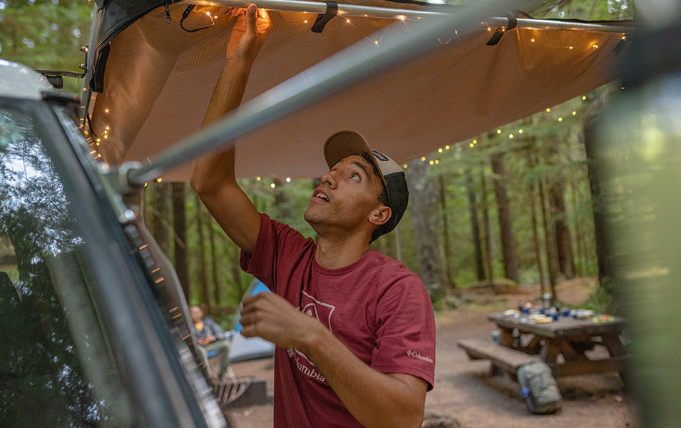A man adjusts camping lighting on the back of his vehicle in a scenic forest campsite. 