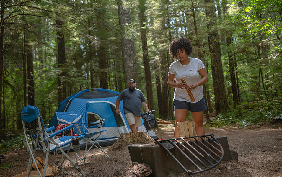  A woman gets a campfire started in the forest as a man walks in front of the tent behind her. 
