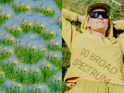 A woman lounges in the grass under bright sunlight, wearing a light green Sunshield™ Hoody with the phrase "GO BROAD SPECTRUM" cast in shadow across the front. She pairs it with a matching cap and shorts, along with oversized sunglasses. Beside her, a prismatic view of a field of yellow dandelions creates a dreamy, kaleidoscopic visual effect.