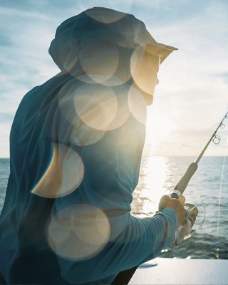 A fisherman in a sun-blocking hoodie photographed from behind. He's on a boat and the sun is shining creating lens flare.