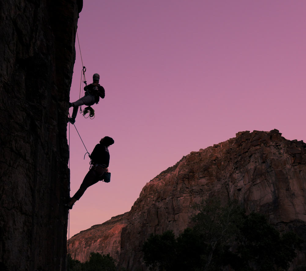 Sillhoutte of one climber and one photographer taking a photo of them, rappelling above, while outside during a sunset, climbing in the desert.