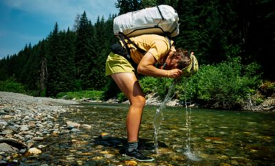 Hannah takes a sip of water from the river, hunched over with her pack.







