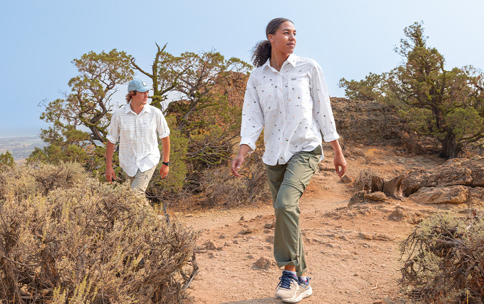 A woman wearing a UPF-rated Columbia Sportswear Silver Ridge shirt hikes through a high desert landscape with a man in UPF clothes trekking behind her. 