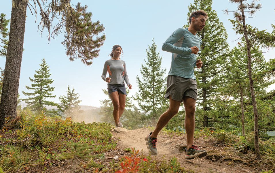 A trail runner wearing UPF-rated clothing runs down a path with a woman jogging behind him. 
