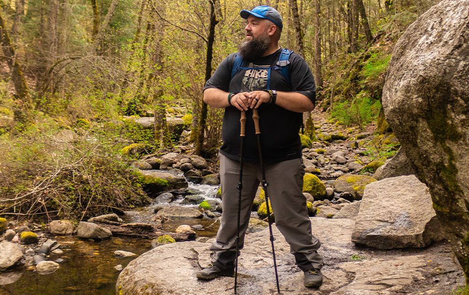 Andy Neal, an outdoor filmmaker and host of The Hiker Podcast, stands in a scenic wooded area holding a pair of hiking poles.