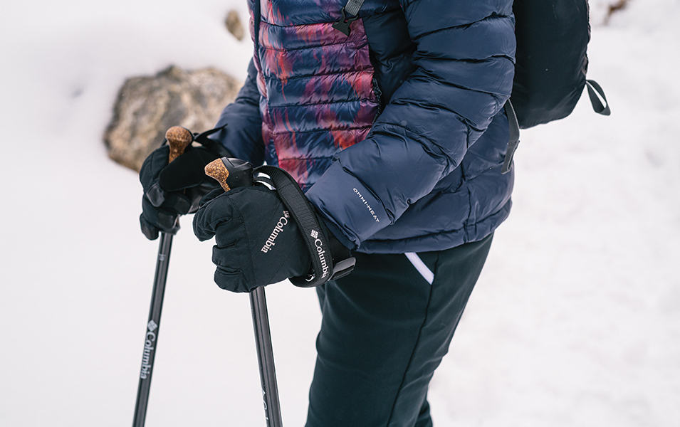 A woman in a blue jacket holds a pair of Columbia Sportswear trekking poles amid a snowy winter background.