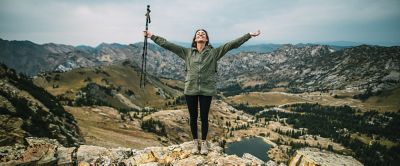 A woman in a green jacket stands on top of a mountainous landscape with her arms in the air holding a pair of trekking poles.
