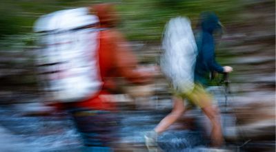 Abstract motion blur image of hikers crossing a stream, wearing large backpacks and outdoor gear. The photo captures dynamic movement with streaks of color, emphasizing speed, activity, and the energy of backcountry adventure.