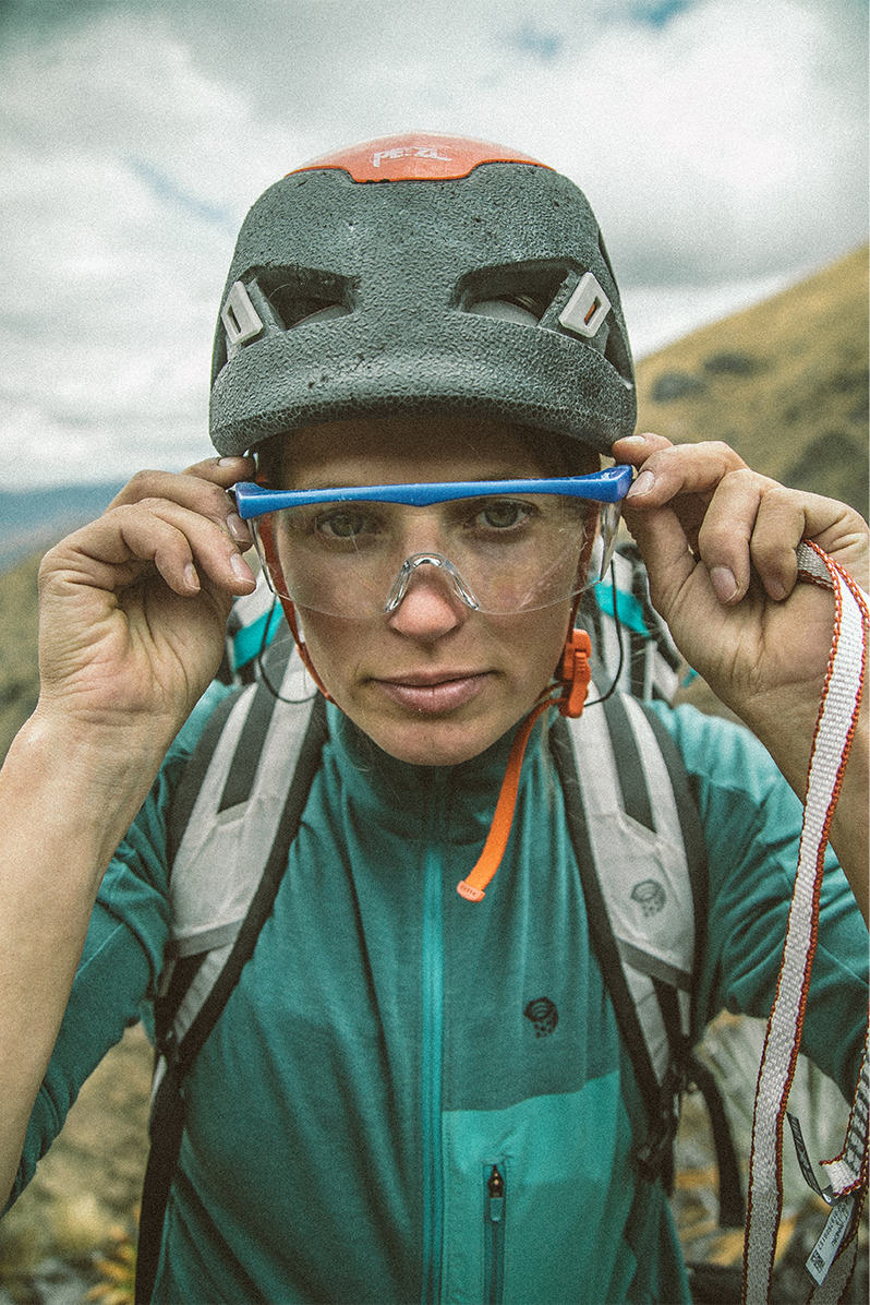 Up close profile shot of Charlotte, looking into the camera lens with climbing helmet and protective eyewear on, gears up for the next route setting.