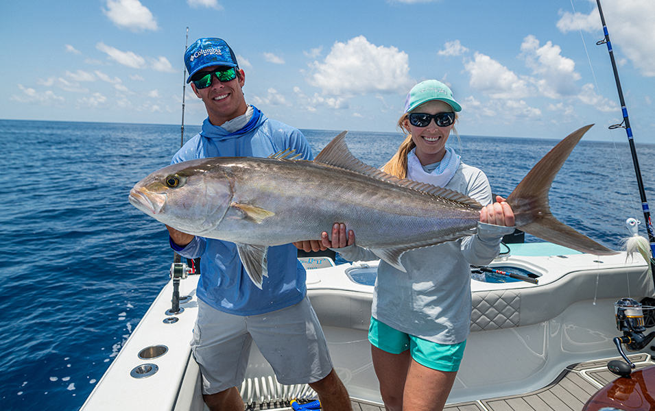 A young man in a blue long-sleeved Columbia Sportswear Terminal Tackle shirt holds up a giant fish on a boat next to a young woman wearing a similar long-sleeved white shirt. They are both wearing baseball caps and smiling.  