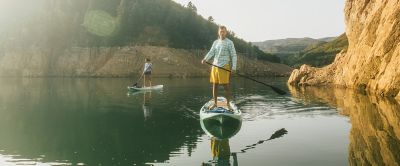 A young man in a light blue long sleeved shirt paddleboards in a scenic area.