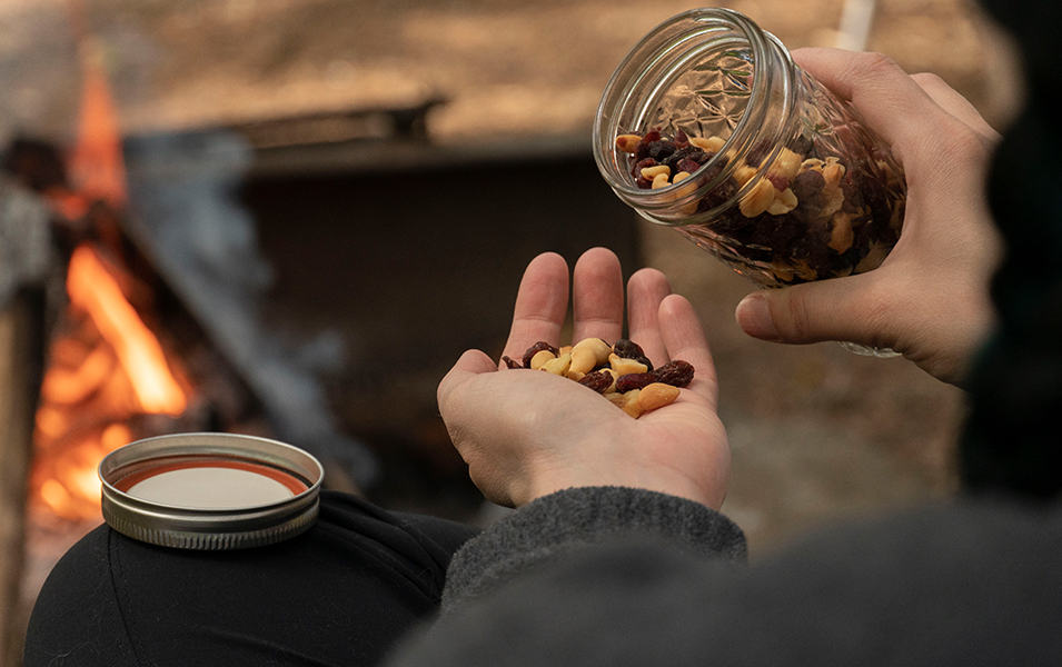 Person pouring out snack mix from glass jar near campfire. 