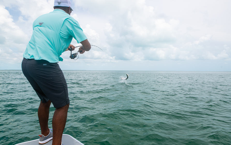 A man wearing an aqua-blue Columbia Sportswear fishing shirt and navy-colored shorts reels a fish into his boat from the ocean.