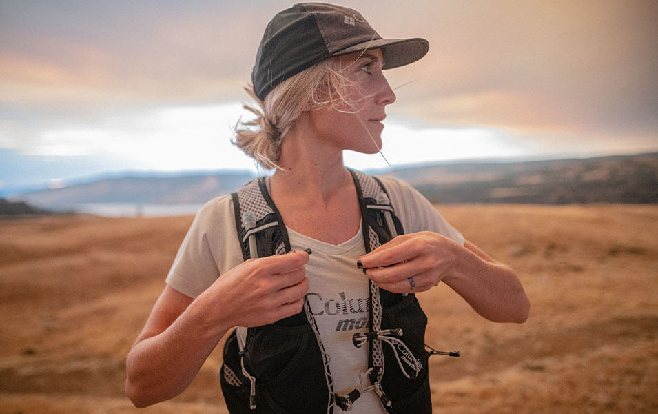 A trail runner adjusts her hydration vest while looking off camera in a scenic outdoor setting. 
