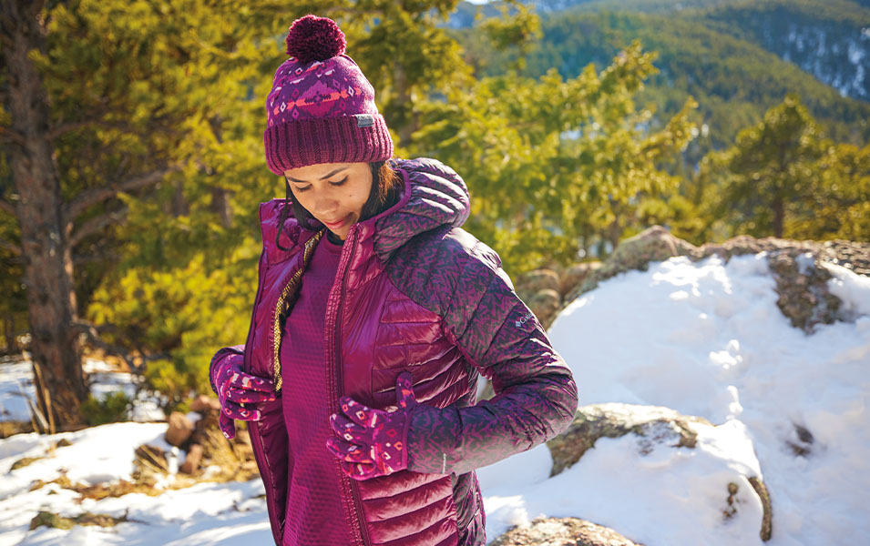 Une femme portant un chandail en laine polaire et un manteau matelassé est debout sur un sentier de randonnée en hiver.