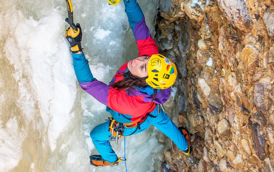 Pro climber Nikki Smith scales a vertical rock face covered in ice while wearing a bright multi-colored jacket and yellow helmet.