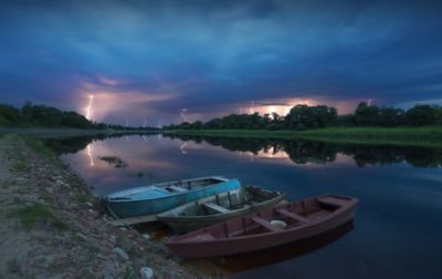 A narrow inlet with three row boats pulled up to rocky ground is threatened by a magnificent storm. The dark and cloudy sky is illuminated by more than a dozen lightning bolts. 