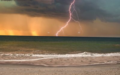 A big thunderstorm crashes down over an ocean. In the forefront are waves and a sandy beach; in the background, there are black clouds. A jagged bolt of lightning strikes the water as the sun sets to the west.  