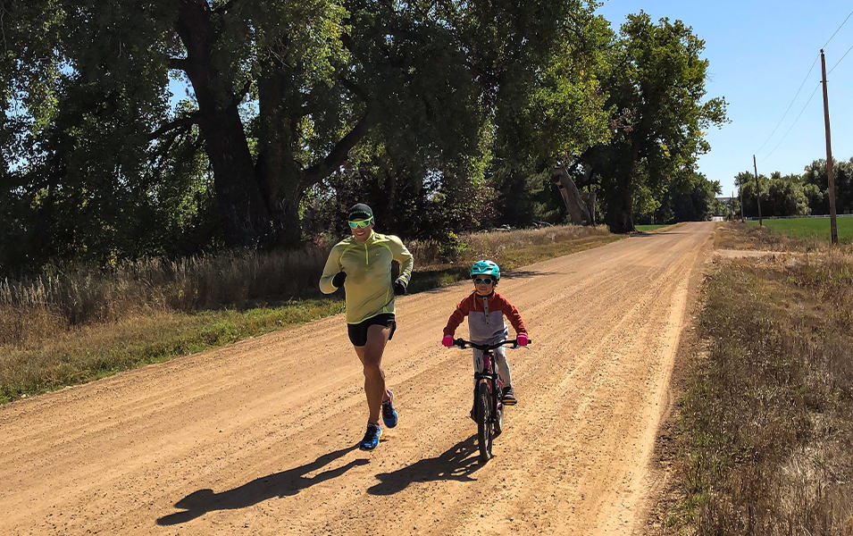 Man running next to child riding a bike on a dirt road. 