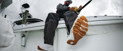 A man sits on the rail of a boat while reeling in his line; the grippy, textured soles of his PFG Pro Sport Boots are visible 