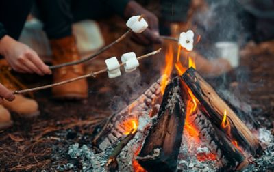 Campers toast marshmallows around a tipi-style campfire with glowing embers.