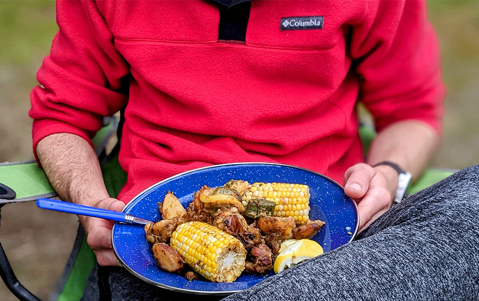 A man in a red Columbia Sportswear jacket sits in a camping chair outside holding a blue dish full of grilled food.