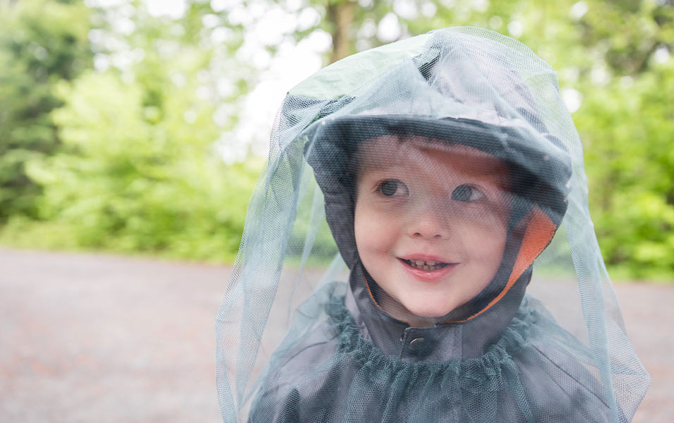 A boy stands outside wearing mosquito netting with green shrubbery in the background.
